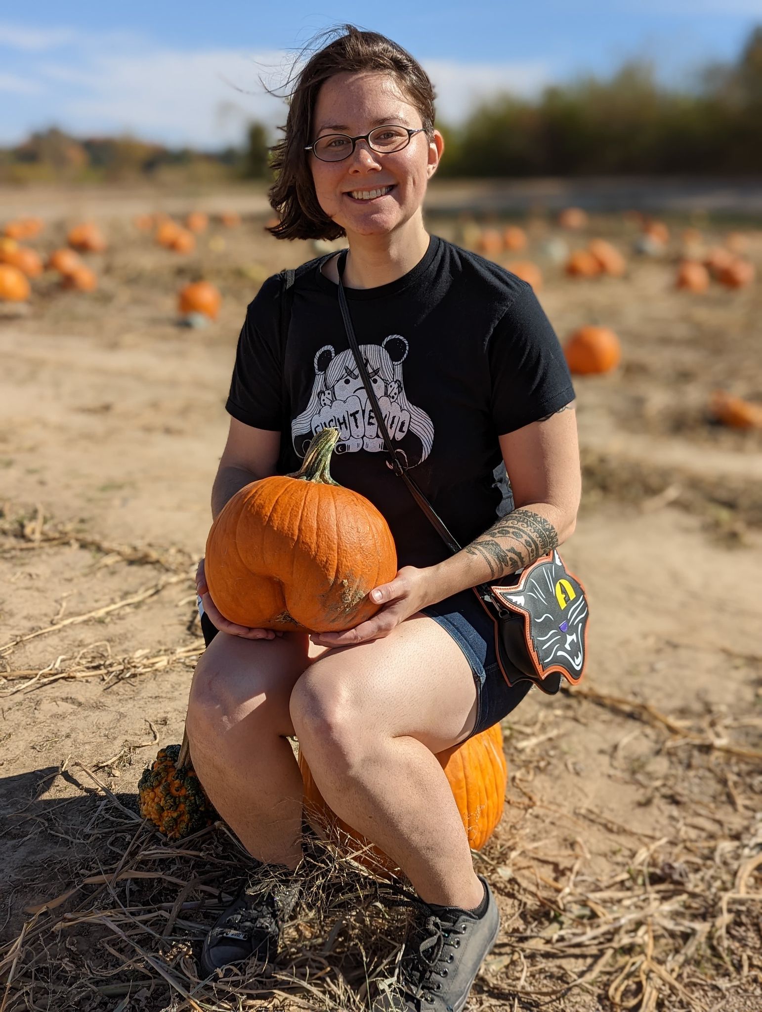 A white woman with windswept brown hair, sitting on a pumpkin in the middle of a pumpkin field. She is holding a pumpkin with a crevasse that makes it look like it has a butt or, arguably, like a character from Among Us. She looks immensely pleased.